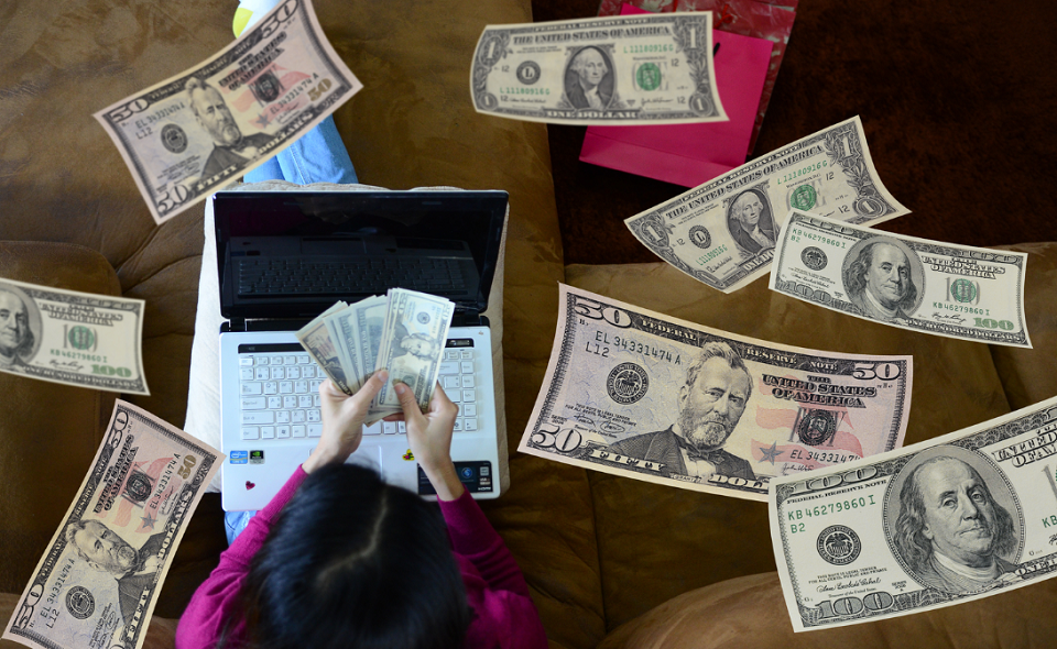 woman using laptop, holding cash, with cash floating down